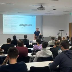 Referent vor einer Präsentationsfolie mit Dow-Jones-Logo, davor Studierende an Tischen in einem Seminarraum.